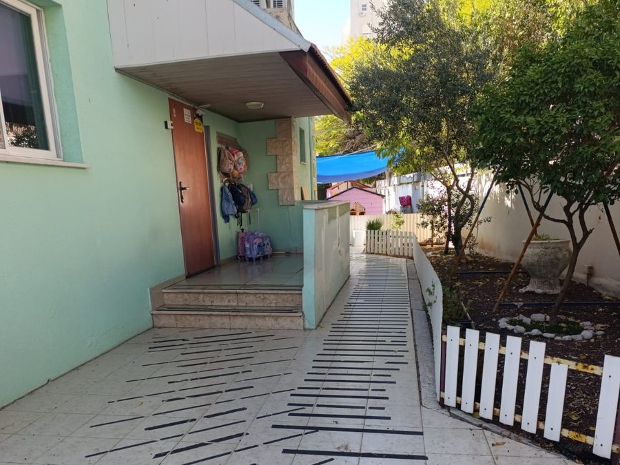 הכניסה לגן — רמפה עם פסים שחורים, דלת חומה וגדר לבנה, בבניין בצבע מנטה